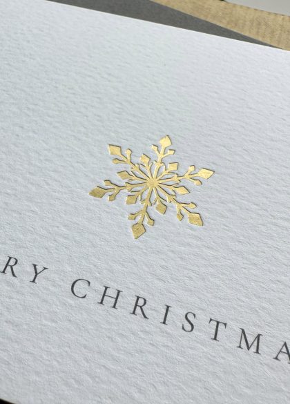 Luxury gold foil snowflake Christmas card on textured white paper with grey envelope and festive wrapping.