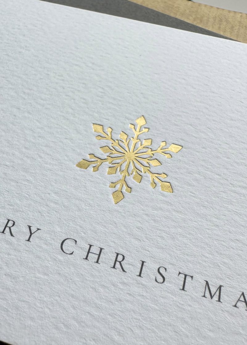 Luxury gold foil snowflake Christmas card on textured white paper with grey envelope and festive wrapping.