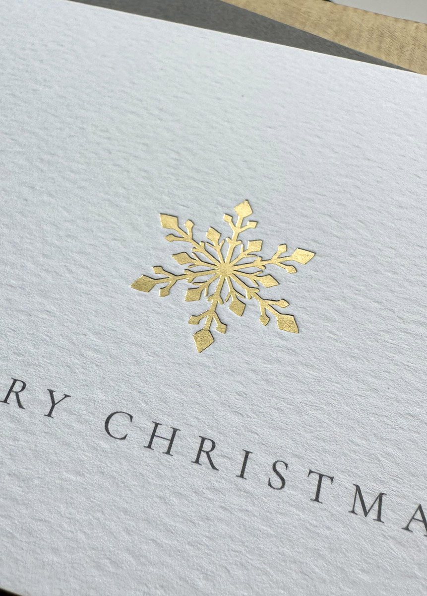 Luxury gold foil snowflake Christmas card on textured white paper with grey envelope and festive wrapping.