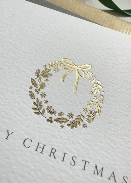 Luxury Merry Christmas card featuring close up of a gold foil wreath design, displayed on a wrapped gift with holiday decorations.
