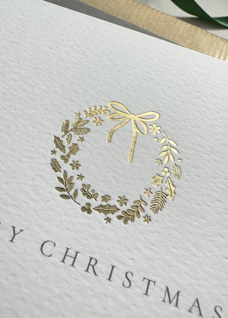 Luxury Merry Christmas card featuring close up of a gold foil wreath design, displayed on a wrapped gift with holiday decorations.