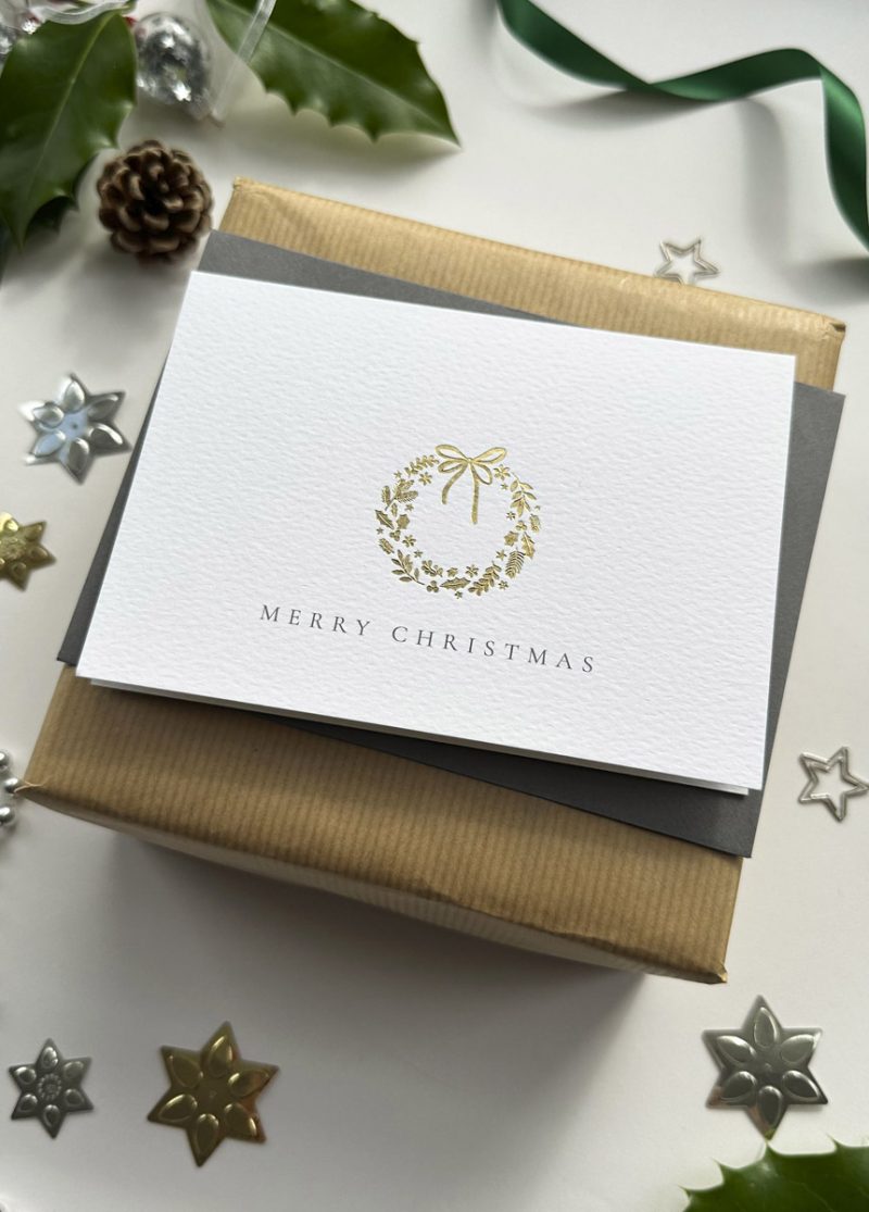 Elegant gold foil Christmas wreath card on luxury textured paper with grey envelope and festive gift wrap.