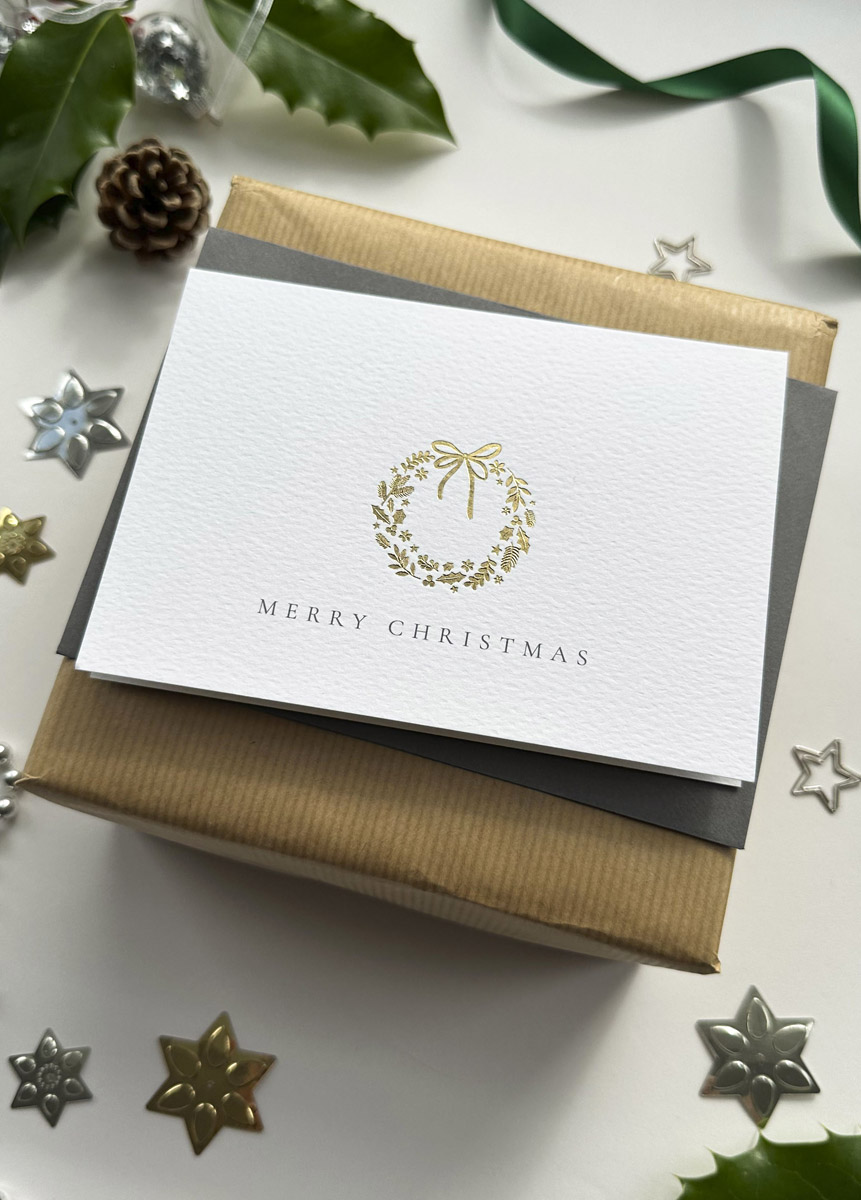 Elegant gold foil Christmas wreath card on luxury textured paper with grey envelope and festive gift wrap.