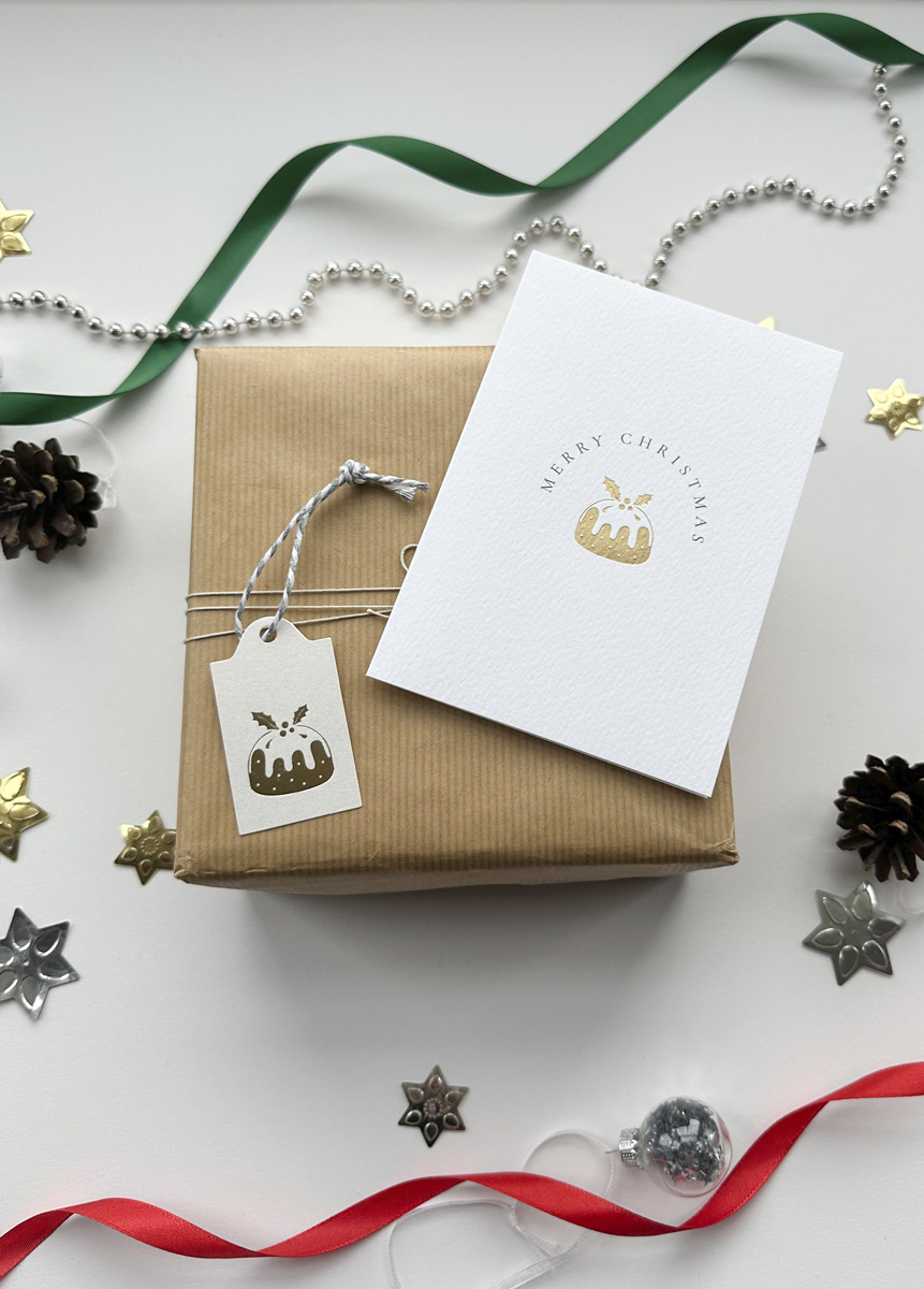 Luxury gold foil Christmas pudding gift tag on kraft wrapped present with festive decorations.